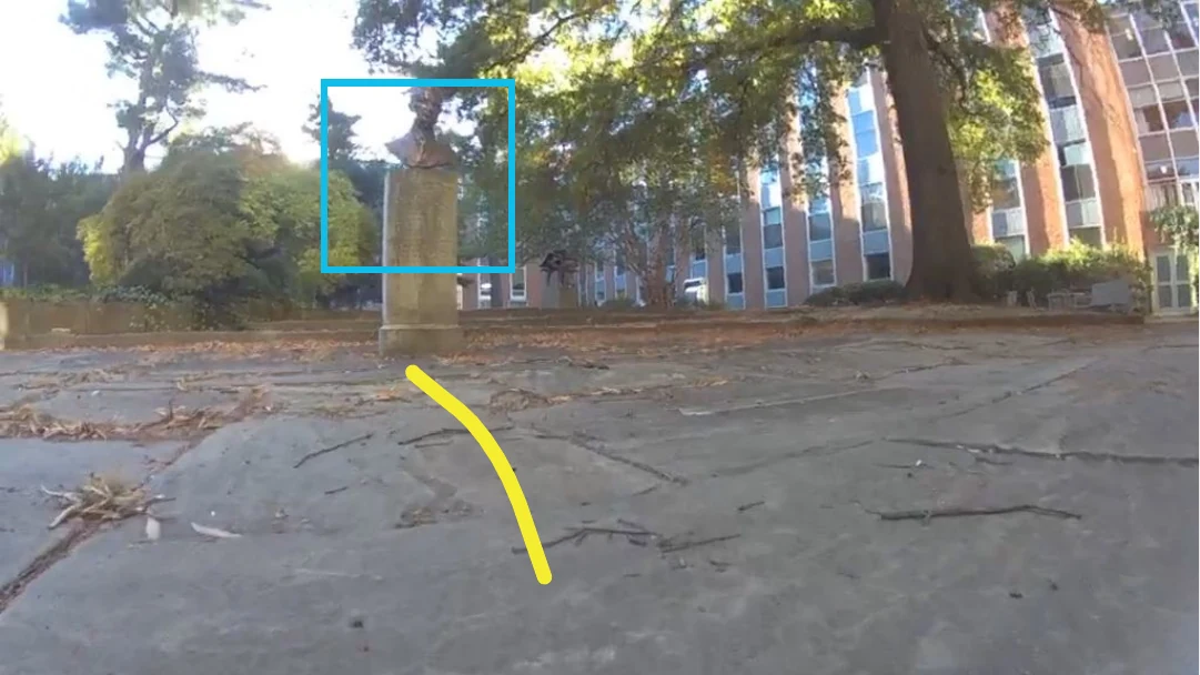 Robot camera view across concrete slabs toward a brick building; a stone statue is boxed on the left and the predicted path curves left toward it.